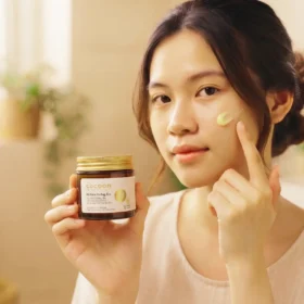 Cô Gái Thoa Hũ Kem Dưỡng Ẩm Cocoon Màu Hổ Phách Lên Má Trong Phòng Tắm Sáng